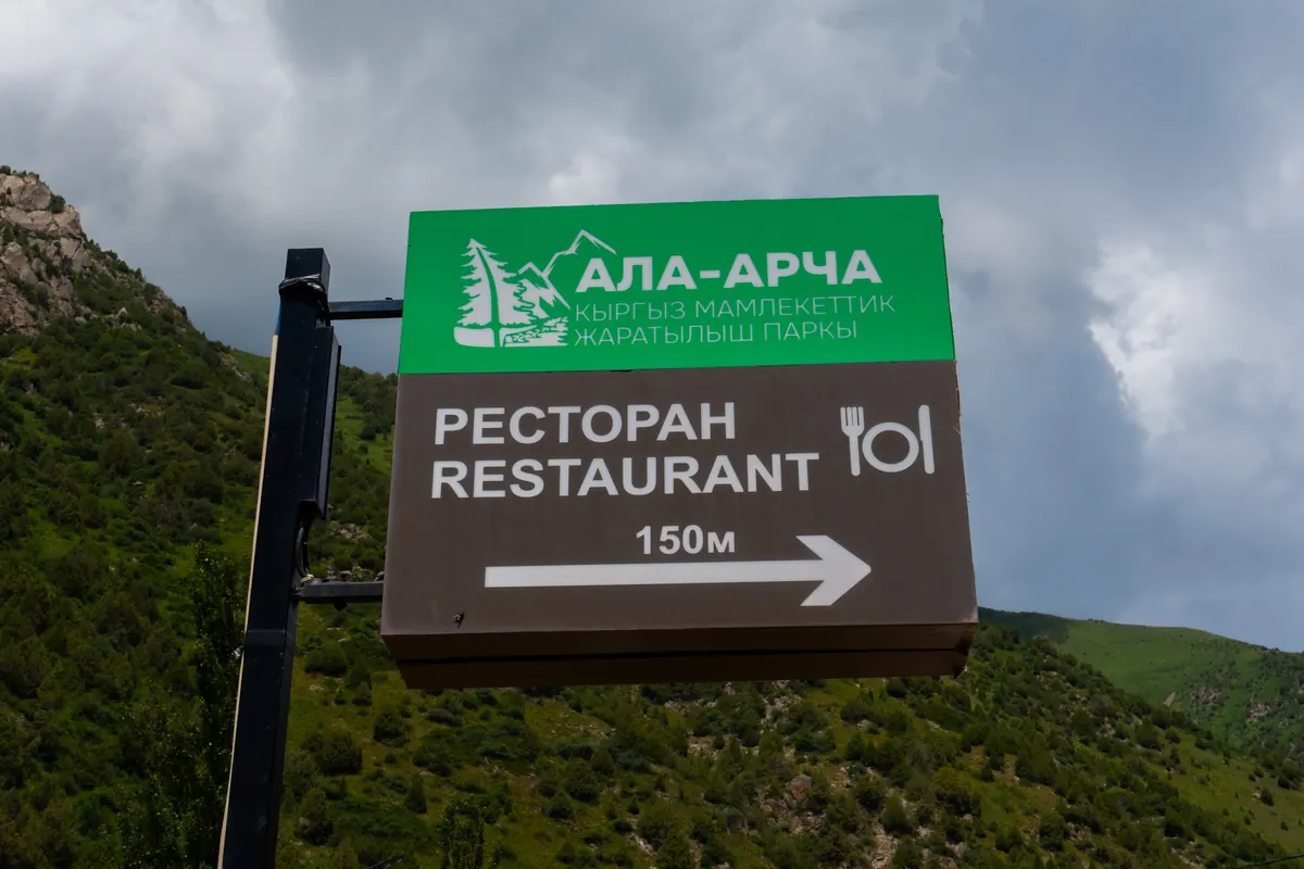 Ала-Арча логотиби бар көрсөткүч: ресторанга 150 м, булуттуу асман жана жашыл беттин фонунда.