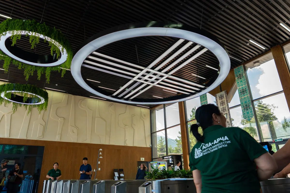 Entrance turnstiles inside the visitor centre under ring lights; a staff member in a green Ala-Archa park shirt is seen from behind.
