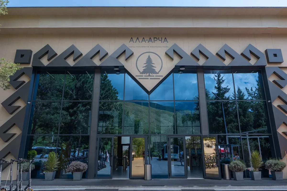 Визит-борбордун маңдайы: кыргыз оюму жана Ала-Арча эмблемасы менен кооздолгон айнек кире бериш, айнекте тоолор менен бактар чагылат.