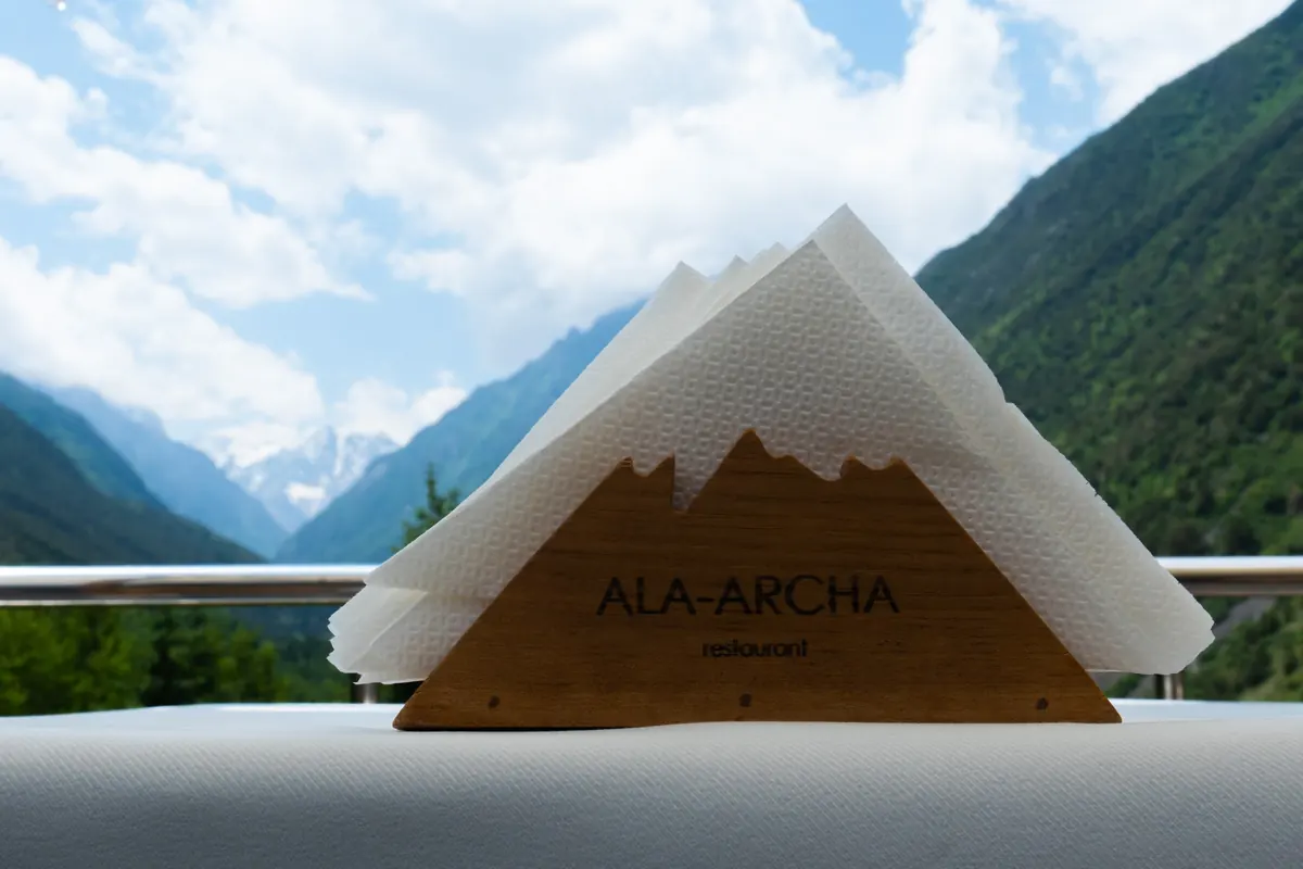 Тоо чокулары формасындагы «Ala-Archa» деп оюлган жыгач салфетка кутусу, ак дасторкондо, артында өрөөн.