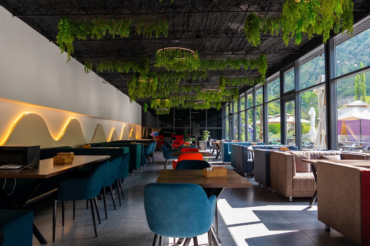 Long restaurant hall with hanging greenery on a dark ceiling, a wavy lit wall, sofas and a row of windows onto the slopes.
