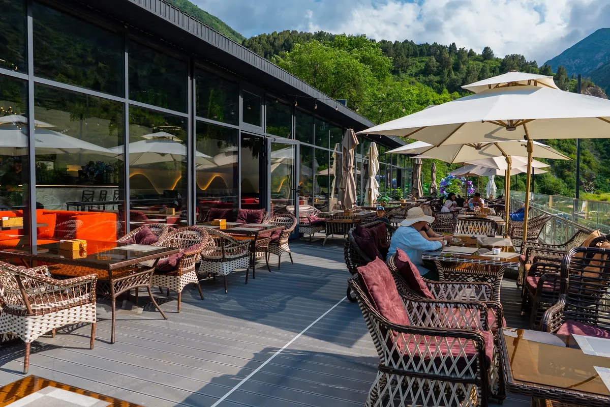 Riverside terrace with wicker chairs, red cushions and cream umbrellas beside a glass-fronted restaurant, wooded slopes behind.