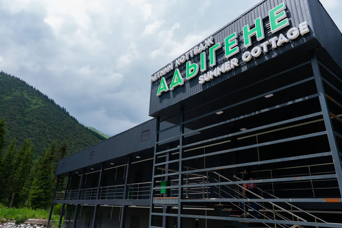 The dark two-storey "Адыгене" summer cottage building with external galleries, green slopes and clouds behind.