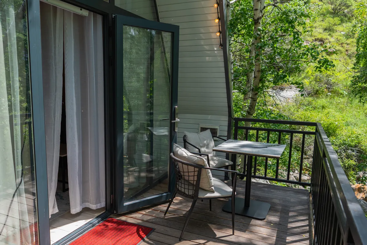 A-frame cabin balcony with a small table and two chairs beside open glass doors, birch trees beyond the railing.