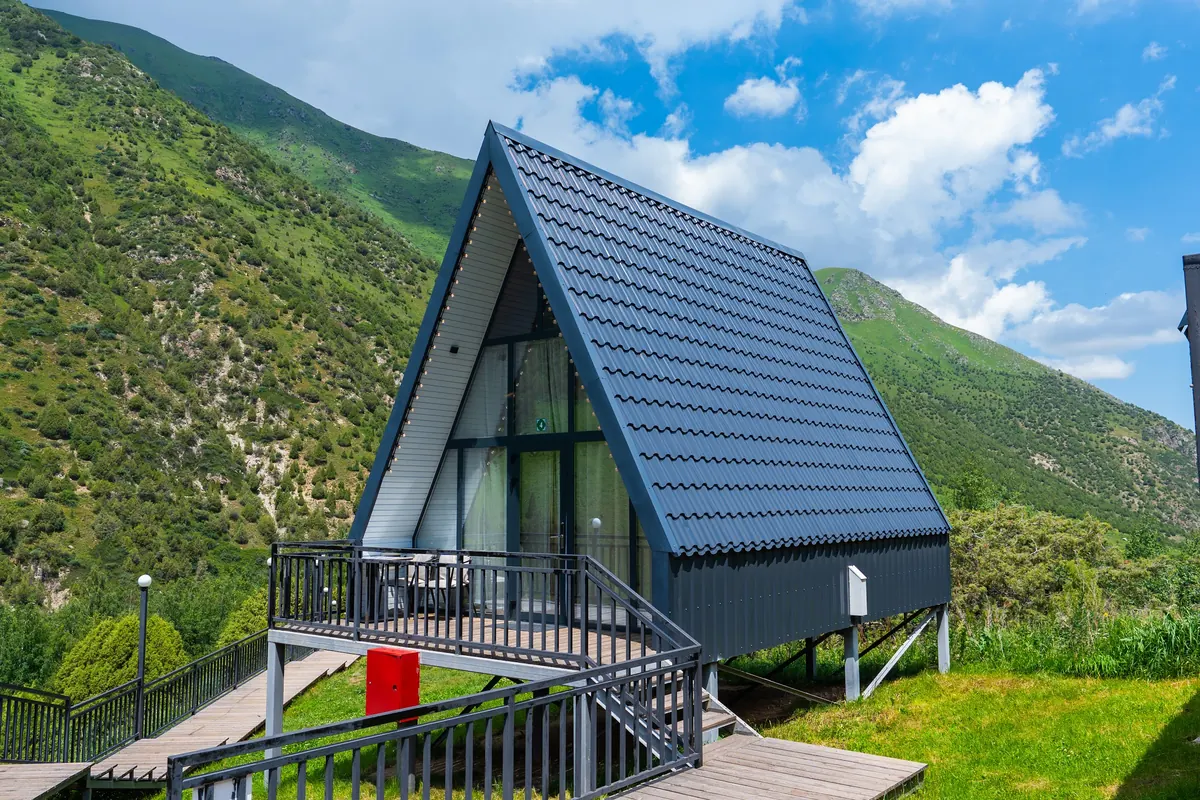 A-frame cabin on stilts with a glass gable and a railed deck, green slopes and blue sky with clouds behind.