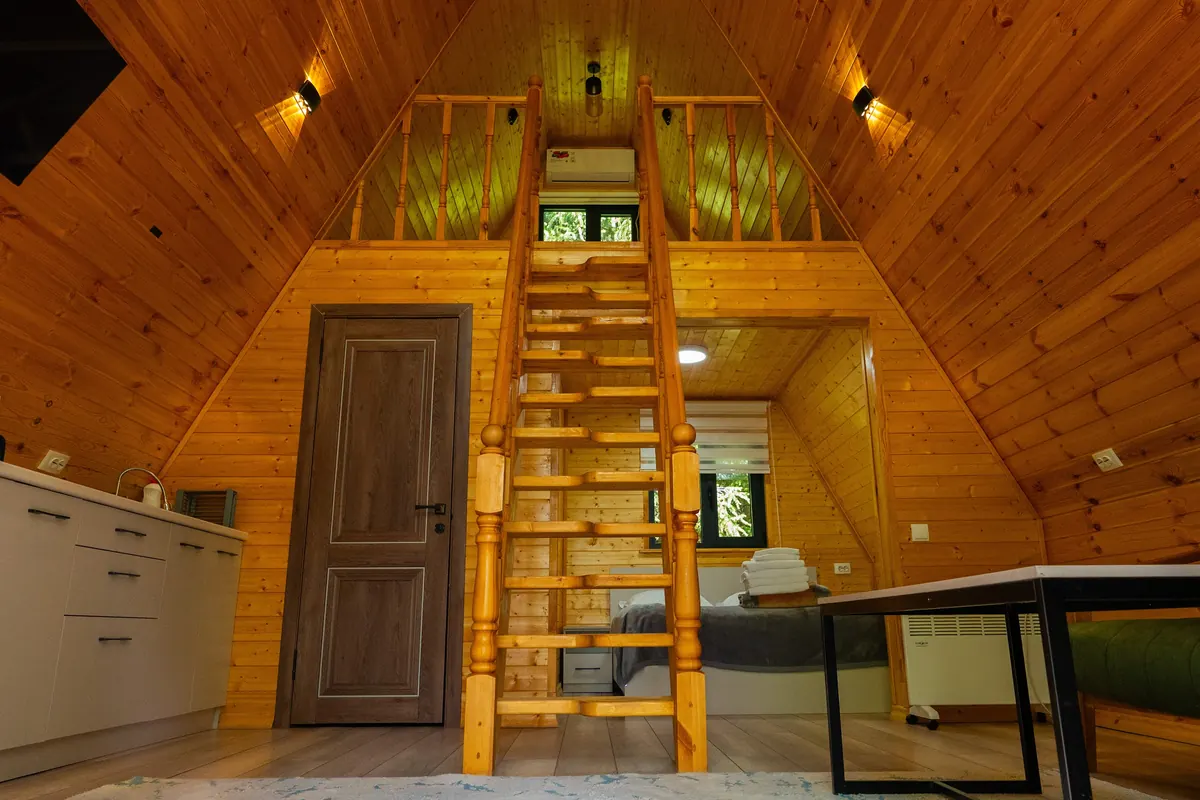 A-frame cabin interior in pine boards: a wooden ladder to the sleeping loft, a kitchenette on the left and a bed in the back room.