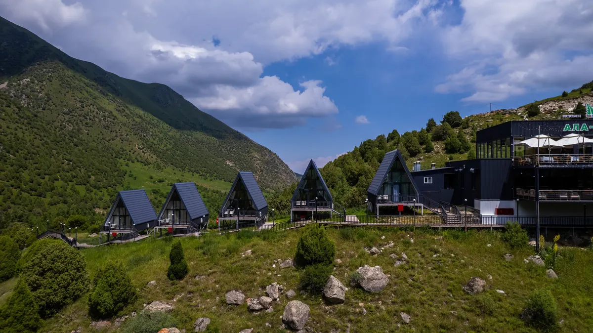 Aerial view of dark A-frame cabins along a boardwalk on a green slope, next to the main restaurant building, with the valley and clouds behind.