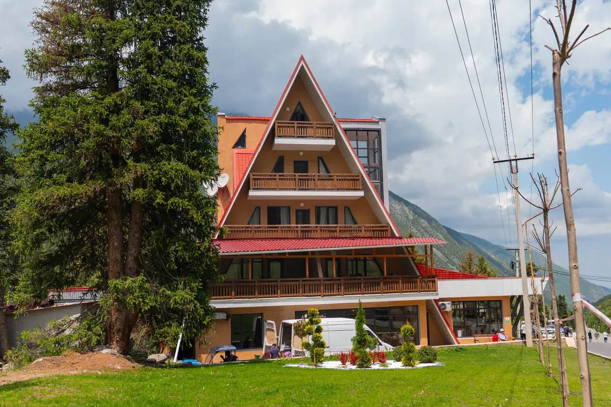 A multi-storey timber chalet with a steep red roof and wooden balconies, beside tall spruces and a lawn.