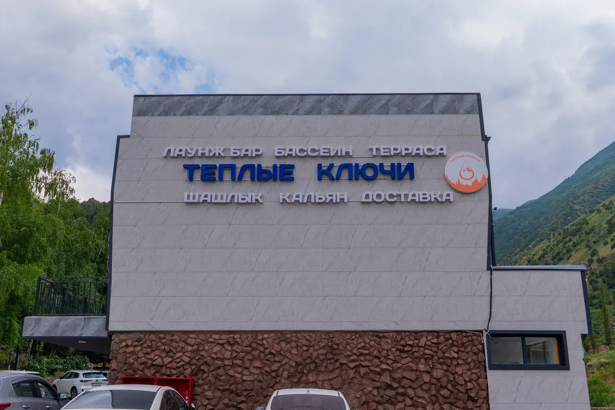 The Teplye Klyuchi (Warm Springs) building with its name in blue letters, on a stone base, wooded slopes behind.