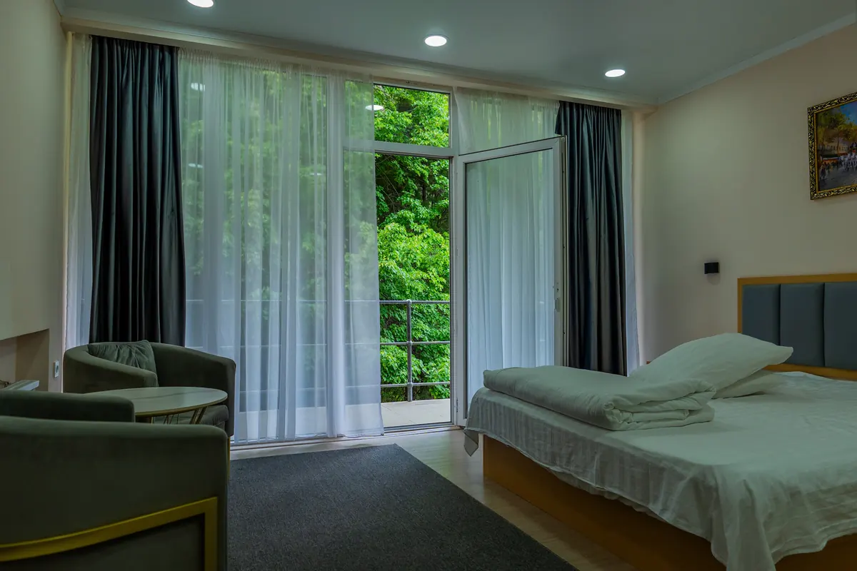 Hotel room with a double bed, two armchairs and a glass balcony door opening onto green trees.