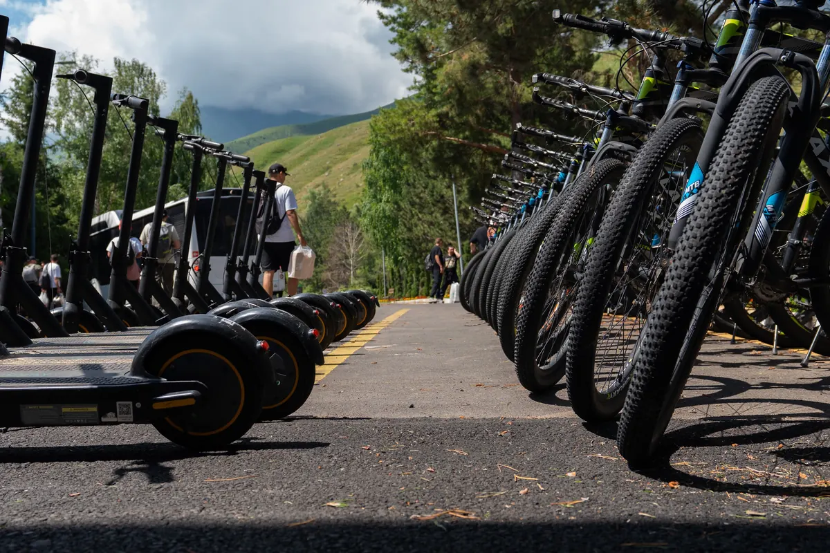 Low-angle view of a rental row of mountain bikes and electric scooters lined up on a path, hills behind.