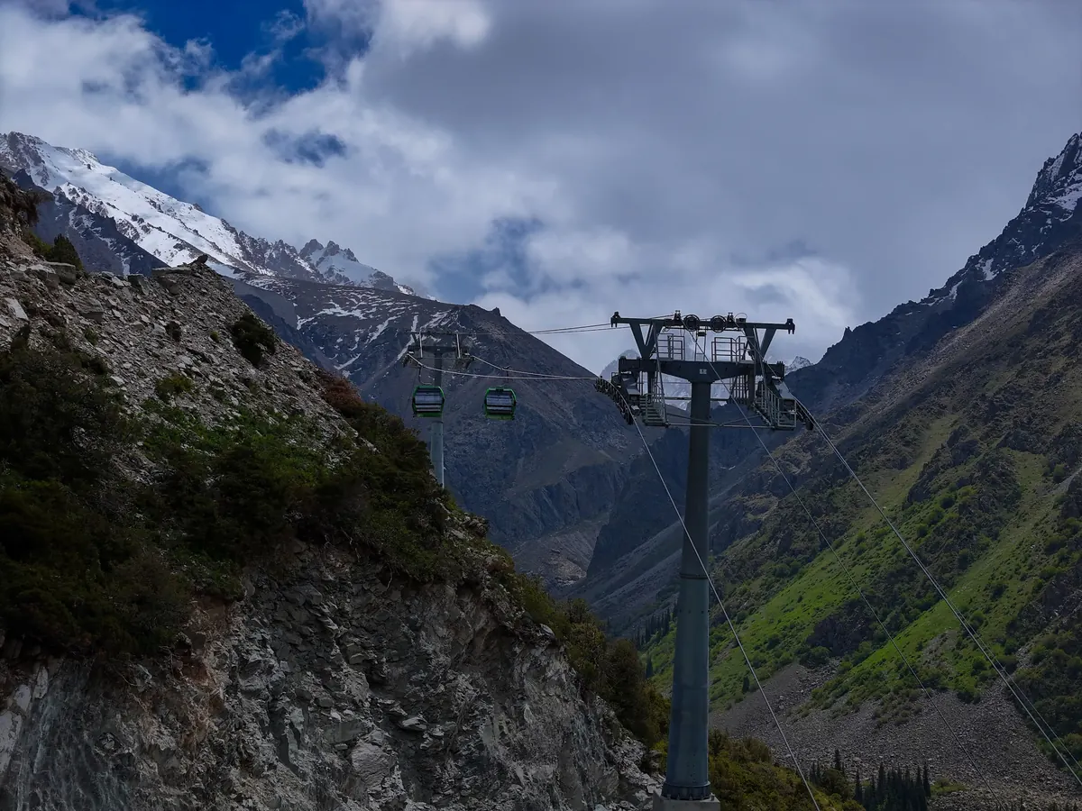 A cable car pylon with two green gondolas on the line, between rocky slopes and snow-dusted peaks under grey clouds.