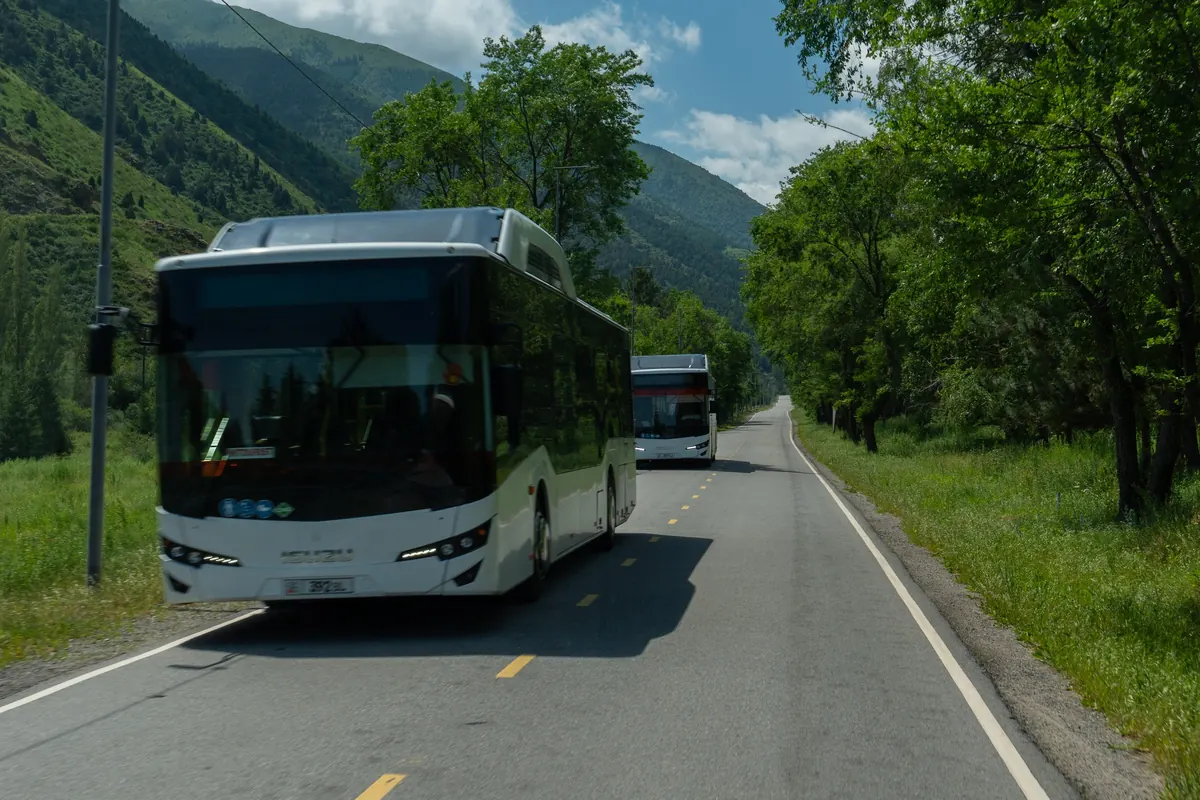 A white electric bus driving along the tree-lined park road, with a second bus behind and mountains beyond.