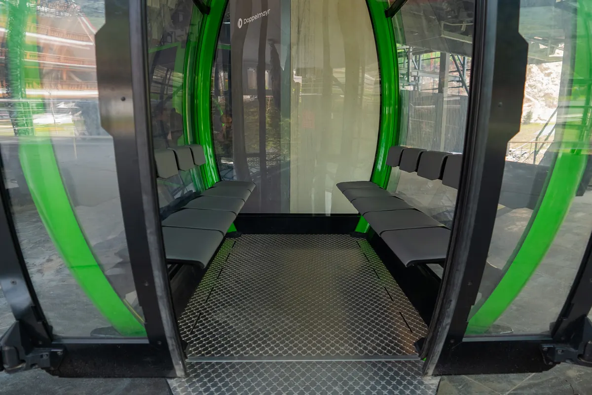 Inside an empty gondola cabin: two facing benches, green frame and large windows.
