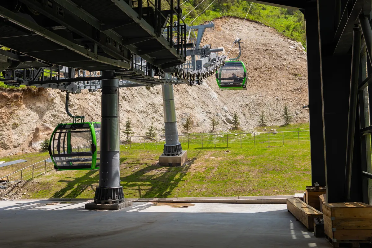 Green gondolas leaving the cable car station and climbing a grassy slope, framed by the station structure.
