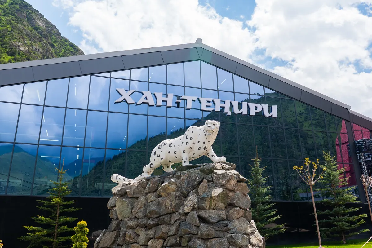Snow leopard statue on a stone base in front of the glass "Хан-Тенири" cable car station, which reflects the mountains.