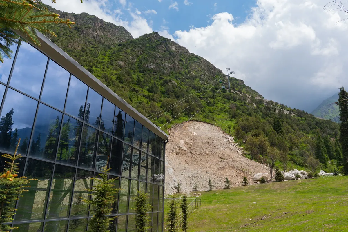 Glass wall of the cable car base station reflecting the slopes, with the cable line climbing a green hillside under clouds.