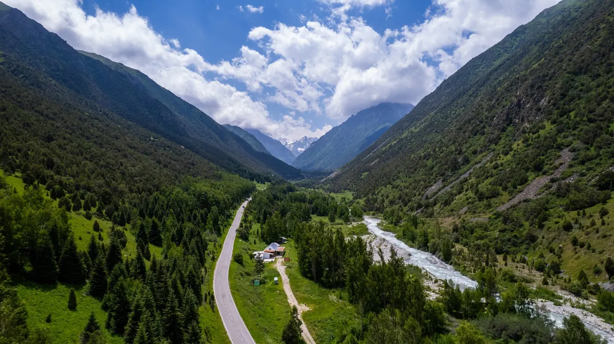 Aerial view up the Ala-Archa gorge: a road and a glacial river run between forested slopes toward snow-capped peaks under white clouds.