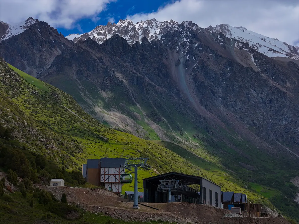 Jagged snow-capped ridge above a green alpine slope, with the upper cable car station and a pylon in the foreground.
