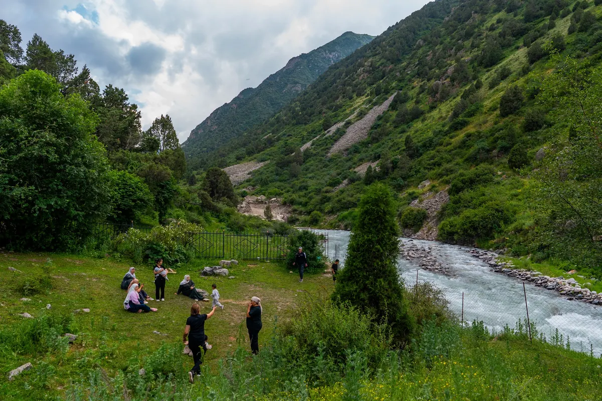 Visitors resting on a grassy meadow beside the river, with forested slopes rising on both sides of the valley.
