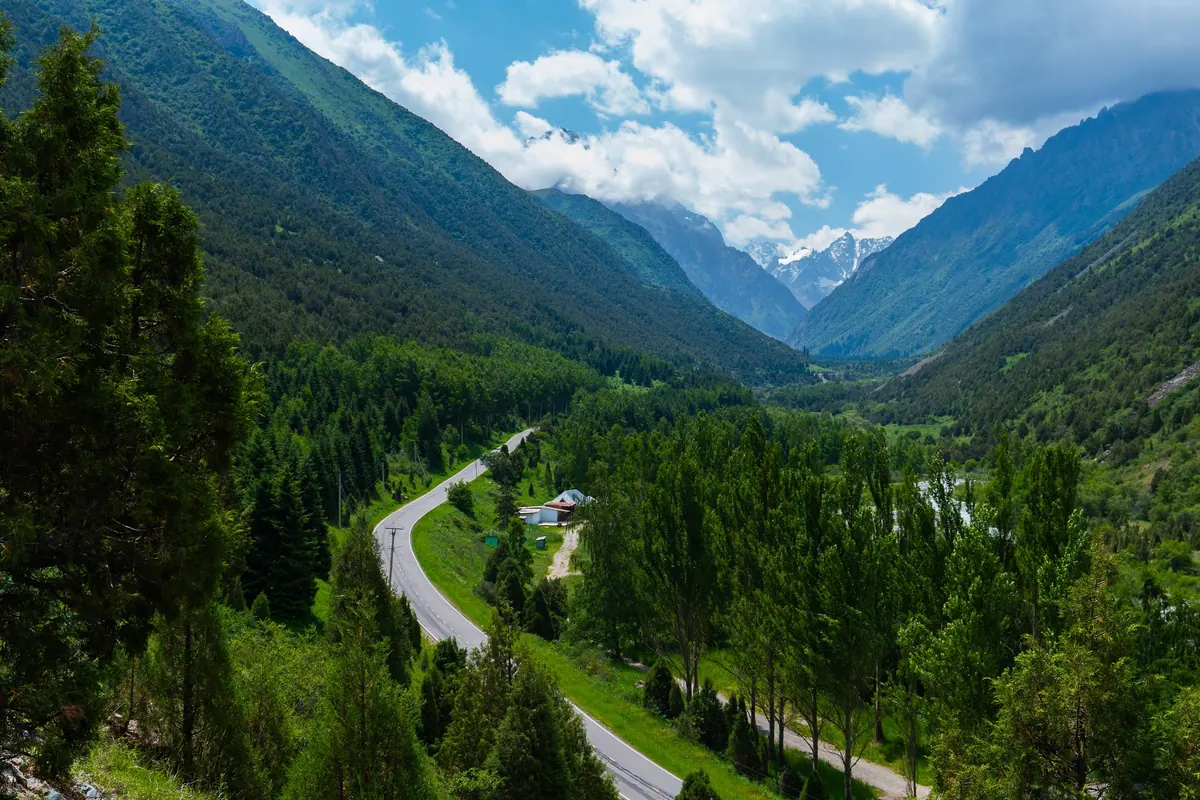 The Ala-Archa valley seen from a hillside: a winding road through conifer forest leads toward snowy peaks under white clouds.